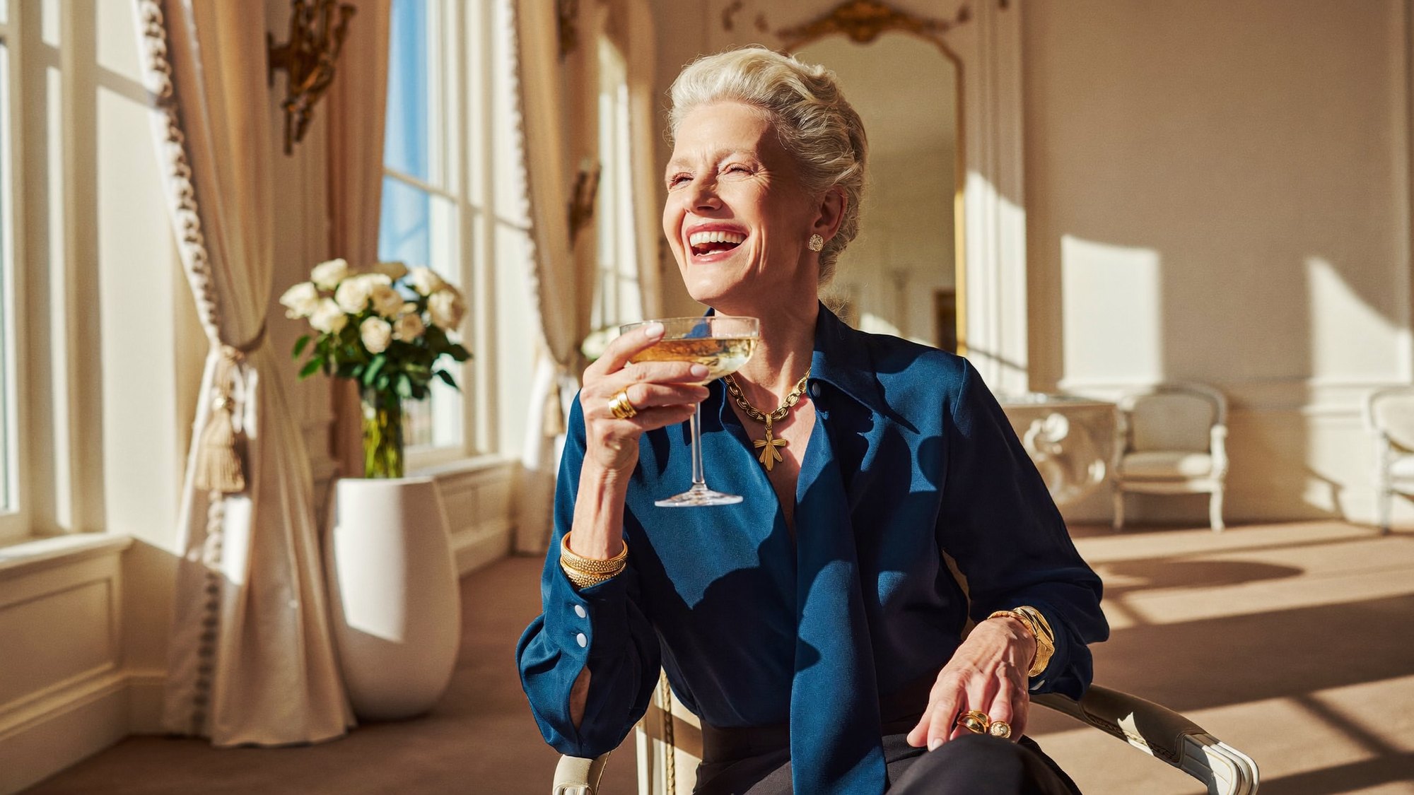 Eine elegante Dame genießt lachend ein Glas Champagner in einem lichtdurchfluteten Salon